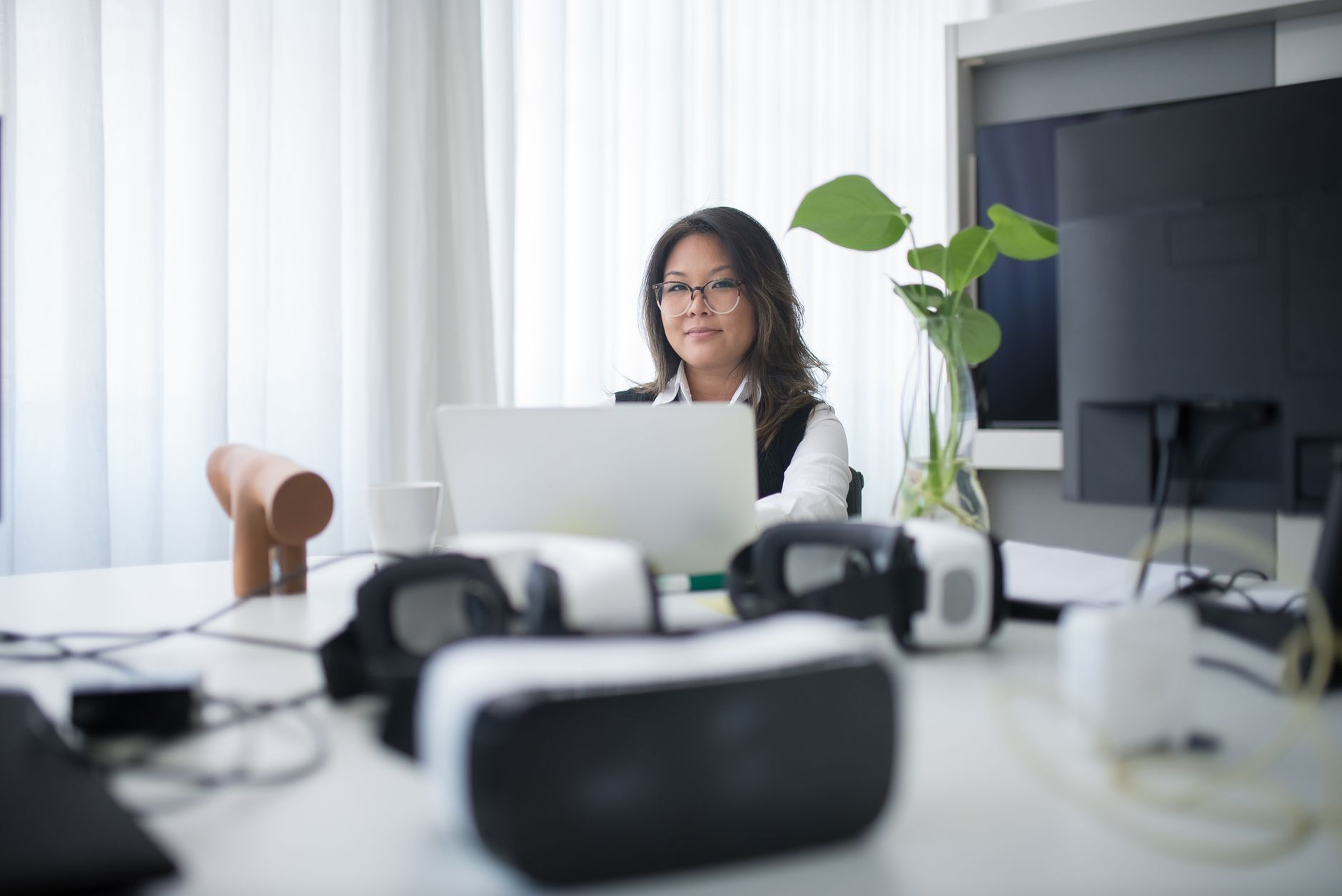 Asian woman in office using laptop with VR headsets on table, symbolizing modern workspace.