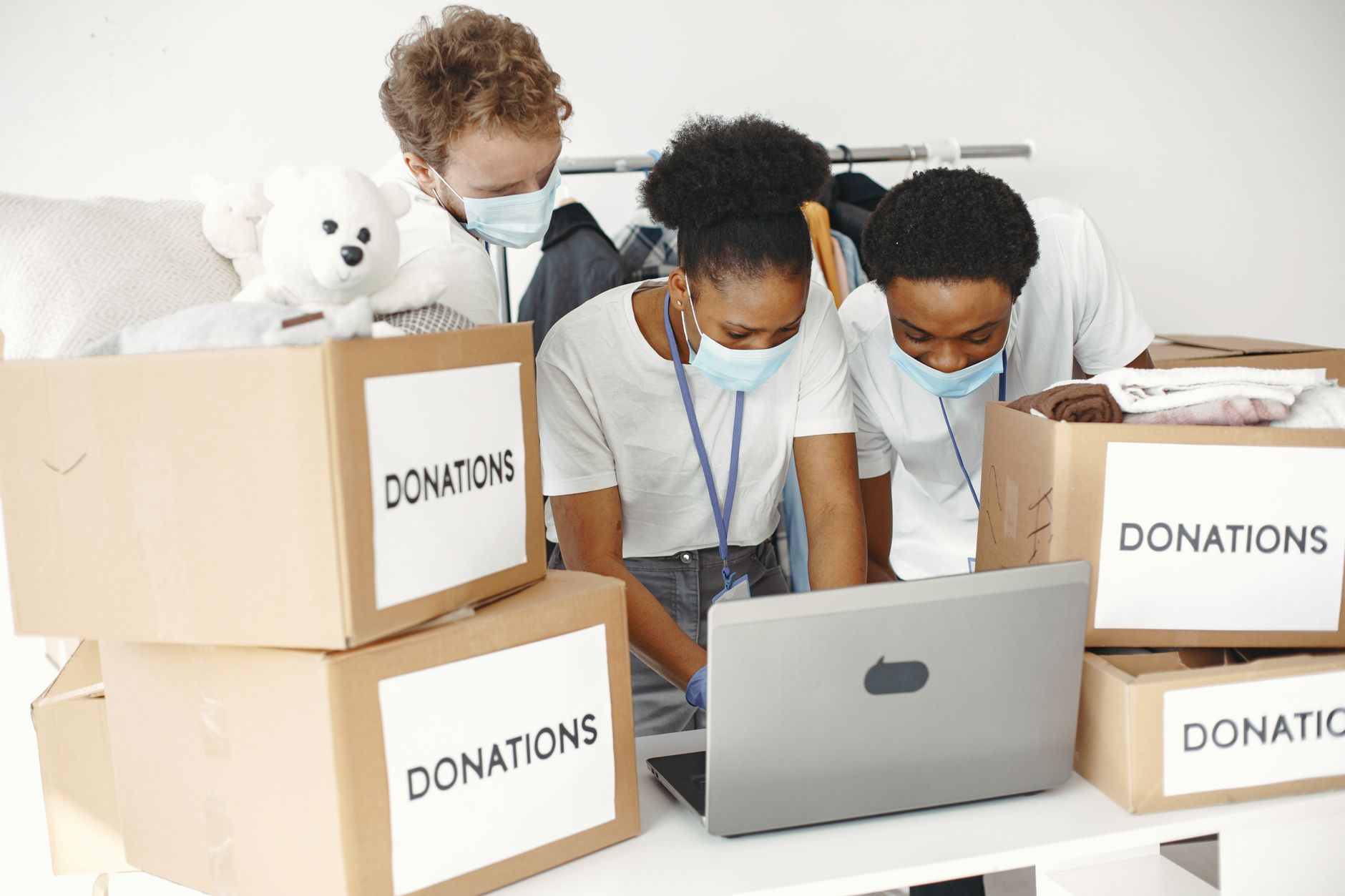 Group of volunteers wearing masks sorting donations with a laptop, promoting diversity and teamwork.