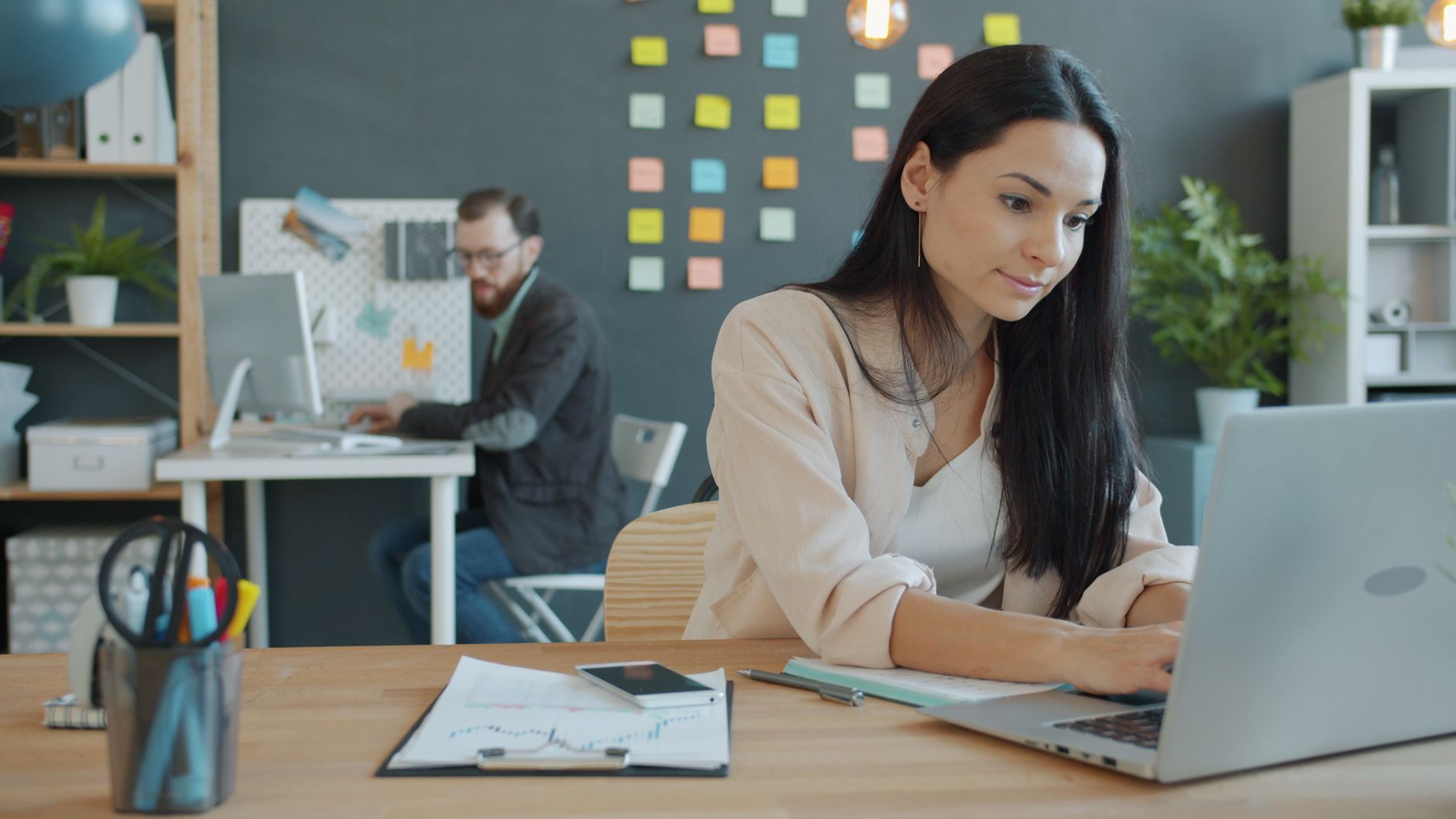 Businesswoman working on laptop in a modern office, with a colleague in the background.